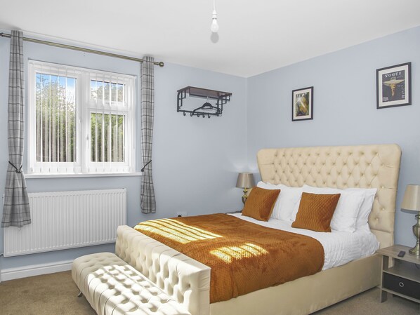 A bedroom with a large bed, a bedside table, a window with curtains, a radiator, and a wall-mounted light fixture.
