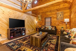A cozy log cabin interior with a large flat-screen TV, a wooden coffee table, a sofa, and a patterned rug.