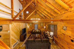 Interior de uma cabana de madeira com teto rústico, um sofá, uma televisão e uma lareira.