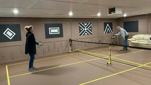En tennisbana med ett nät i mitten, två spelare och en soffa i bakgrunden.