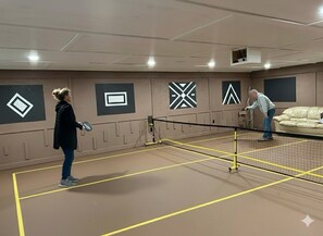 En tennisbane med et net i midten, to spillere og en sofa i baggrunden.