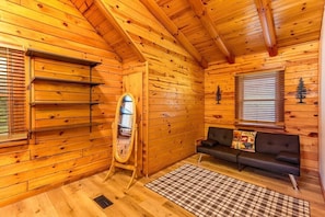 Interior de uma cabana de troncos com piso de madeira, um sofá, um espelho e uma janela com persianas.