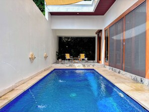 A modern outdoor swimming pool with a clear blue water surface, surrounded by a tiled deck and a covered patio area with chairs and potted plants. - Budayana Villa (Jimbaran)