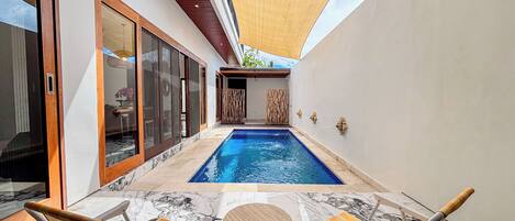 Piscina con dos sillones reclinables y una mesa, rodeada por un muro blanco y un techo de madera.