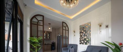 A spacious living area with a modern chandelier, grey sofas, and a kitchen in the background.