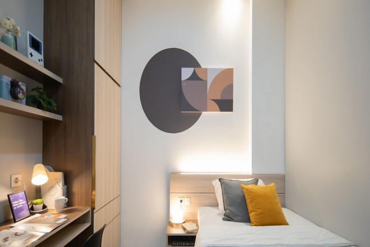 A modern hotel room with a bed, bedside table, lamp, and a wall-mounted abstract art piece.