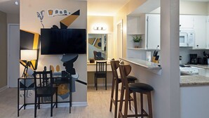 Cocina moderna con armarios blancos, microondas y cafetera. Hay una zona de comedor con mesa y sillas, y un televisor montado en la pared.