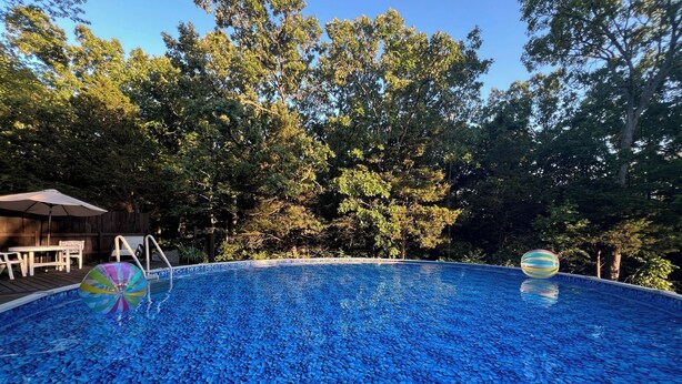 A swimming pool with a blue mosaic tile design, surrounded by trees and a wooden deck with outdoor furniture.