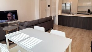 A modern living room with a grey sofa, a white dining table, and a kitchen area with a stainless steel refrigerator.