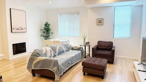 A modern bedroom with a bed, a seating area with a chair and ottoman, a small table with a vase, and a TV.