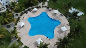 Vista aérea de un resort con una piscina de forma peculiar rodeada de sillas de descanso y sombrillas, palmeras y un edificio con balcones.