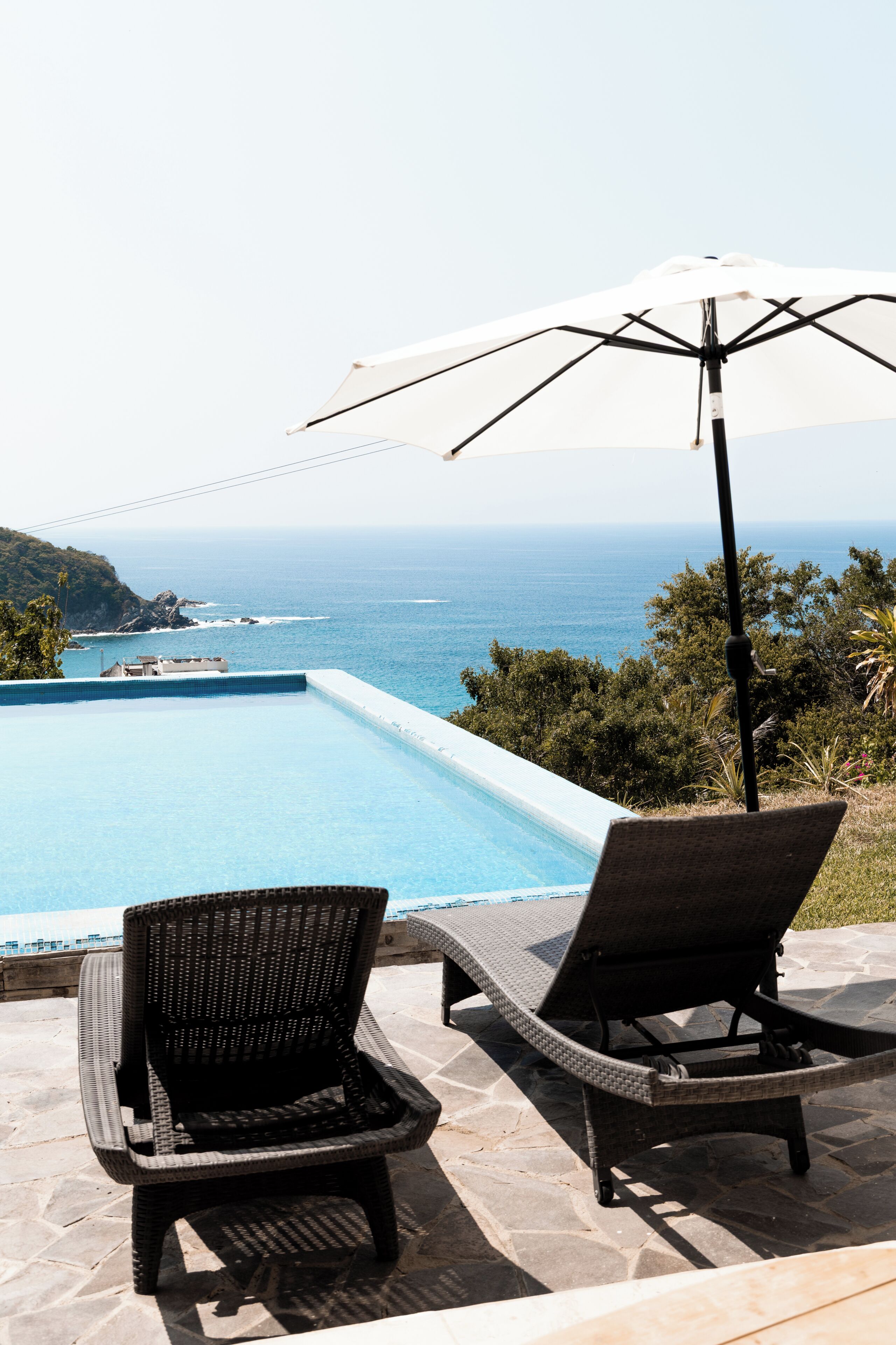 A poolside area with lounge chairs and an umbrella, overlooking a clear blue sea.