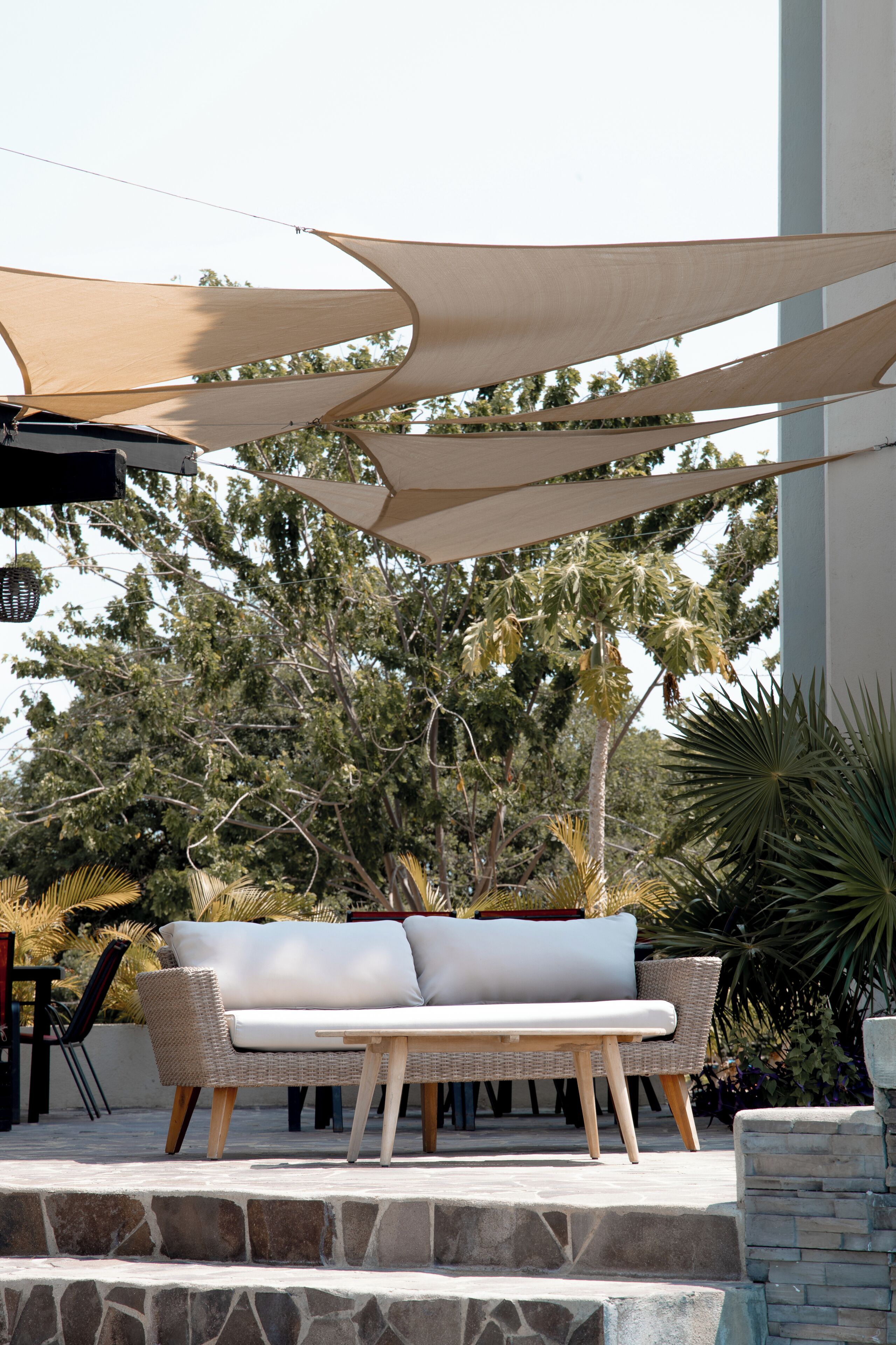 Un área de estar al aire libre con sofá y mesa, protegida por grandes toldos de tela, con árboles y un edificio de fondo.
