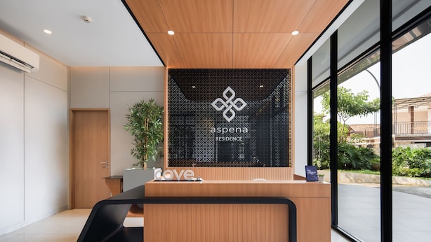 A modern reception desk with a wooden finish, a black mesh partition, and a logo for 'Aspena Residence'. - Cove Aspena Residence (Tangerang)