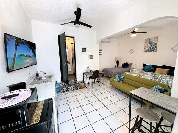 A compact hotel room with a bed, a sofa, a dining table, a TV, and a ceiling fan. - NUEVA VISTA INN (La Cruz de Huanacaxtle)