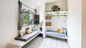 Habitación en esquina con un banco integrado, una ventana con reja metálica y un estante montado en la pared con plantas en macetas.