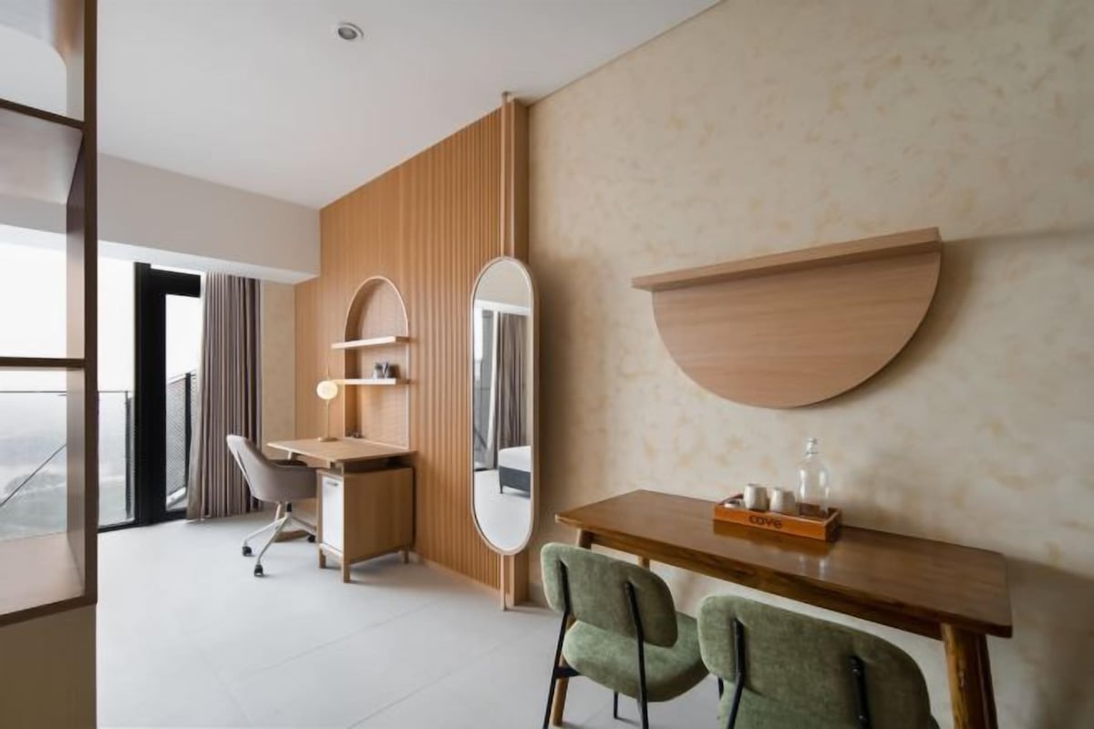 A modern hotel room with a wooden desk, two chairs, a mirror, and a wall-mounted shelf.