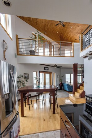 Un'ampia cucina con soffitto in legno, frigorifero in acciaio inossidabile, zona pranzo con sgabelli e una scala che conduce a un piano superiore.