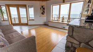 Uma sala de estar espaçosa com piso de madeira, parede de destaque em pedra, uma janela grande com vista para o mar e uma porta de vidro deslizante que dá para uma varanda.
