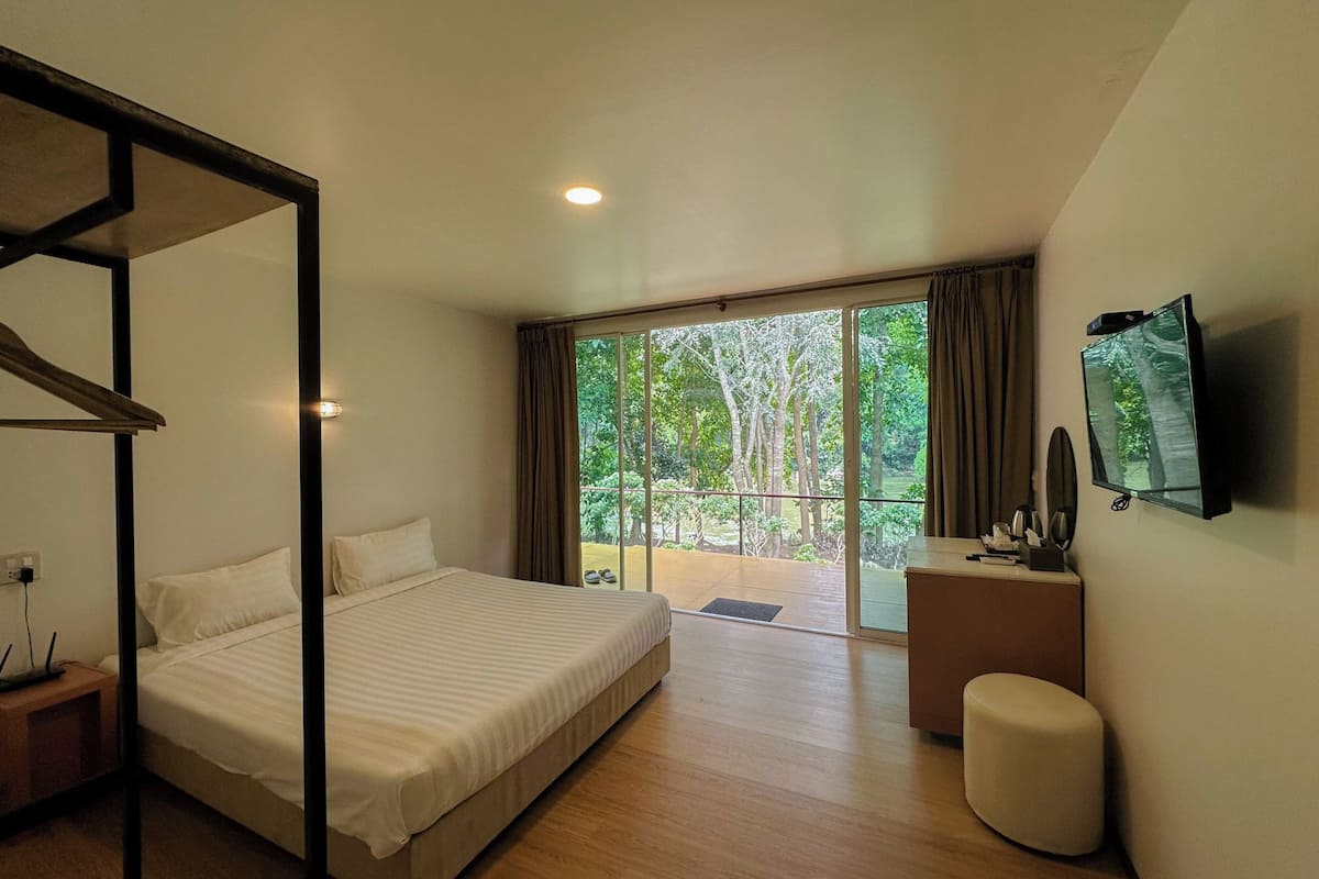Un dormitorio con cama, escritorio, televisor y balcón con vistas a árboles.