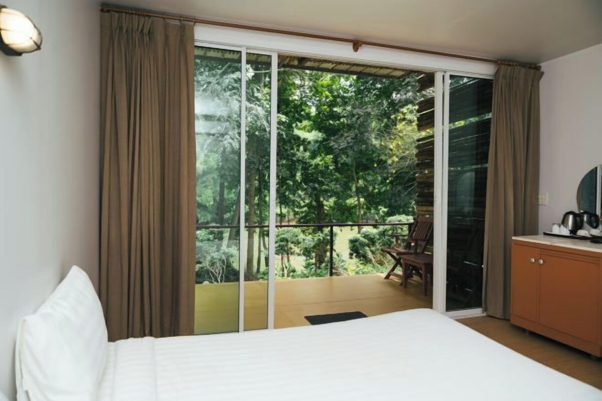 A hotel room with a bed, a sliding glass door leading to a balcony, a small table with a kettle, and a TV on a wooden cabinet.