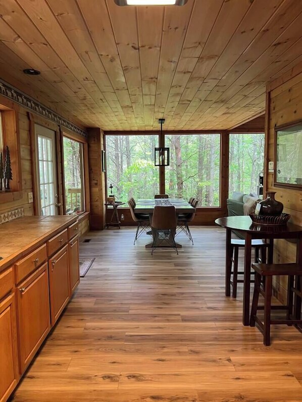 A wooden cabin interior with a dining area, kitchen, and large windows offering a forest view.