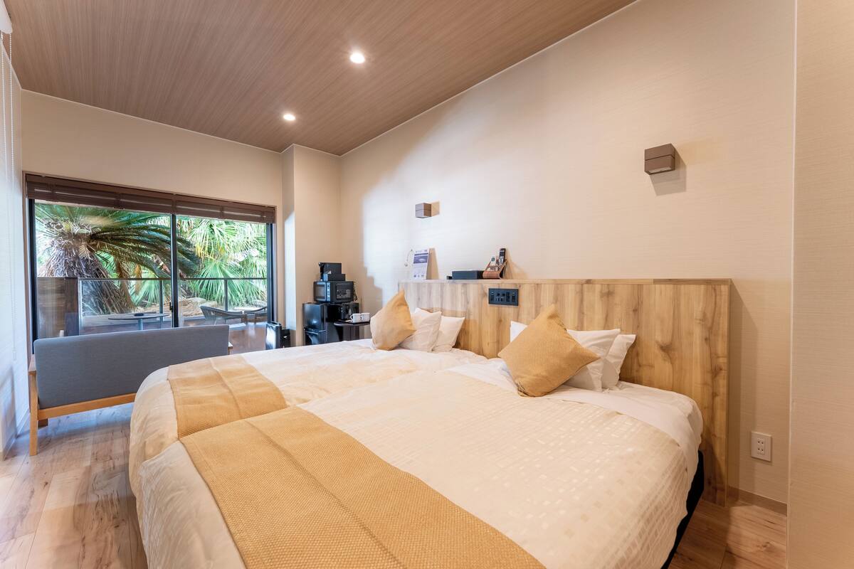 Una moderna camera d'albergo con un ampio letto, una testiera in legno, una scrivania con un computer e vista su palme attraverso ampie finestre.