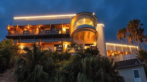 A modern multi-story building with balconies and a curved central section, surrounded by tropical vegetation. - Livemax Resort Inamiseafront (Inami)