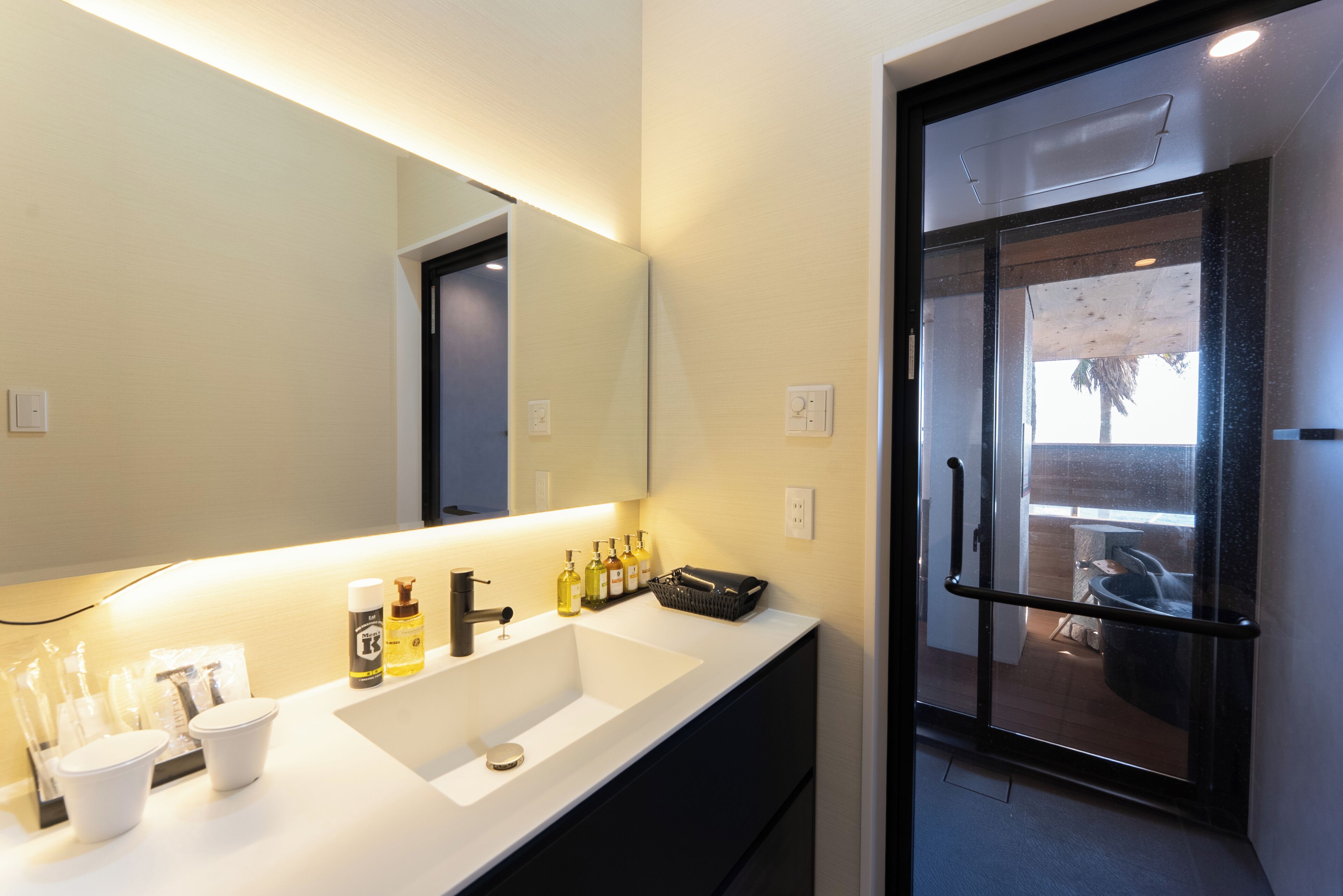 Un bagno moderno con un grande specchio, un lavandino bianco e una porta in vetro che dà su un balcone.