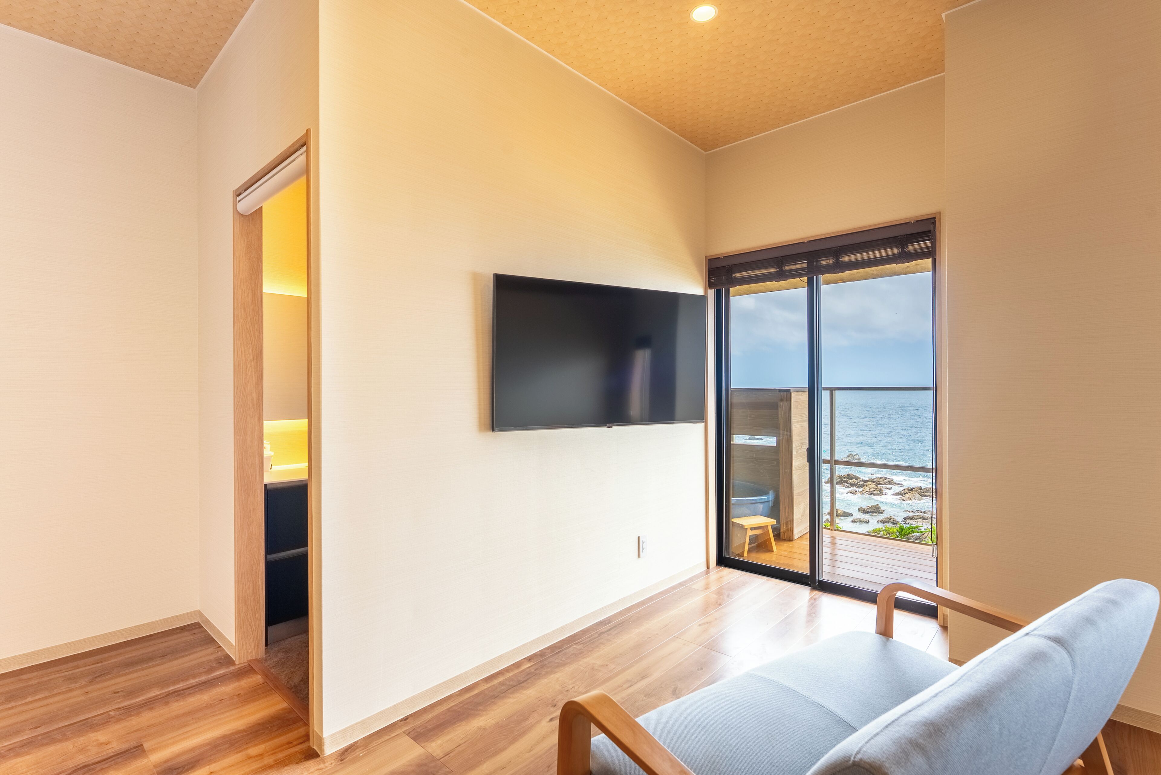 Et moderne værelse med et fladskærms-tv, trægulv, en skydedør til en balkon med havudsigt og en lysegrå sofa.