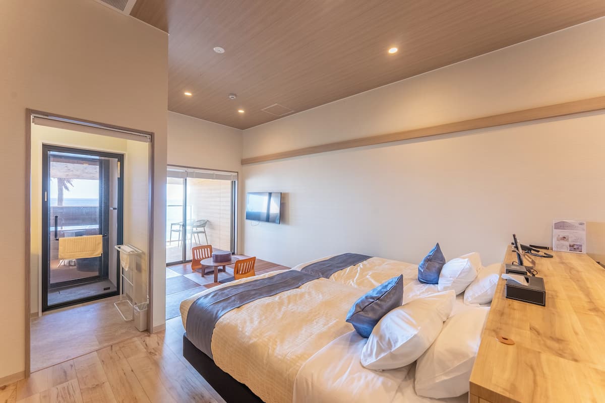 Camera d'albergo moderna con un ampio letto, una scrivania in legno, una TV e una porta-finestra in vetro scorrevole che conduce a un balcone.