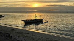Una playa con barcos anclados cerca de la orilla, bajo un cielo nublado con el sol poniéndose en la distancia.