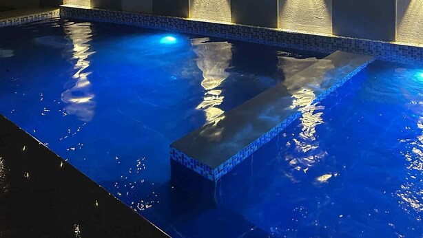 Una piscina con borde de azulejos y pared texturizada al fondo, iluminada por luces colgantes.