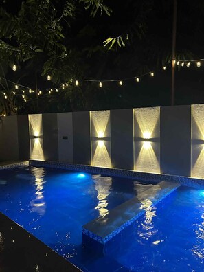 A swimming pool with a tiled edge and a textured wall behind it, illuminated by string lights.
