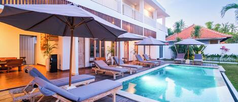 A poolside area with lounge chairs and umbrellas, a wooden deck, and a building with balconies.