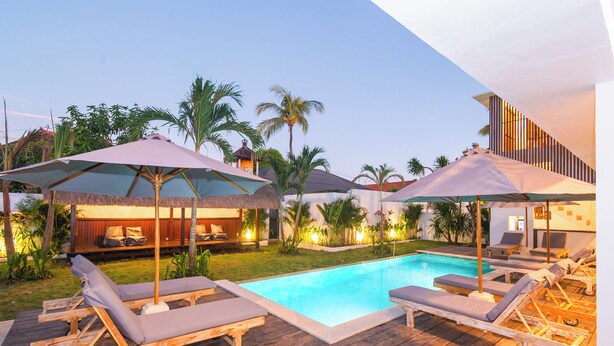 Zona de piscina con sillones de descanso y sombrillas, rodeada de palmeras y una terraza de madera.