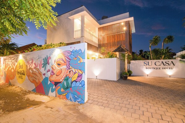 A modern hotel with a colorful mural, a well-lit entrance, and a sign reading "SU CASA BOUTIQUE HOTEL".
