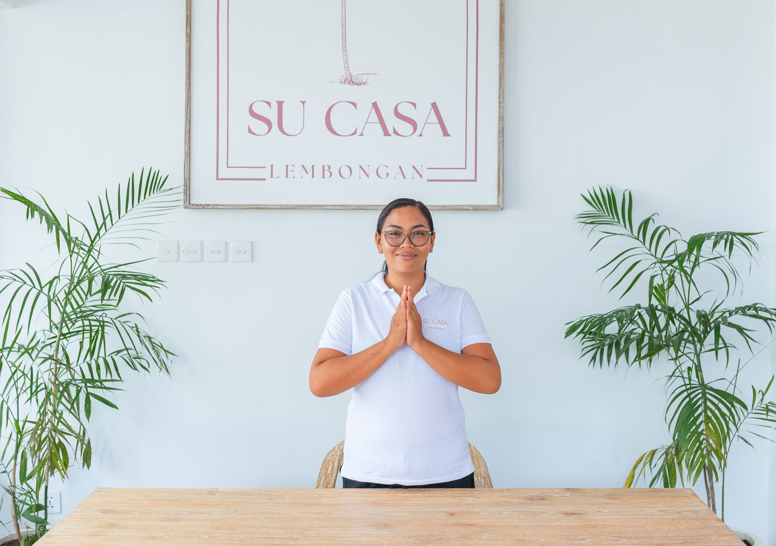 A person in a white shirt with 'SU CASA LEMBONGAN' printed on it, standing with hands together in front of a wooden table and chairs, with two green plants on either side.