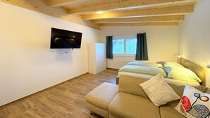 A cozy room with a wooden ceiling, a bed with pillows, a sofa, a TV mounted on the wall, and a window with curtains.
