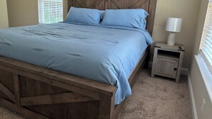 A bedroom with a wooden bed frame, a blue bedspread, and a nightstand with a lamp. - The Red Nest At High Rock Lake - Fire Pit, Front Porch Deck, Sunset & Lake Views (Lexington)
