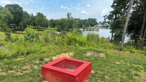 A red fire pit in a grassy area with a lake and trees in the background. - The Red Nest At High Rock Lake - Fire Pit, Front Porch Deck, Sunset & Lake Views (Lexington)