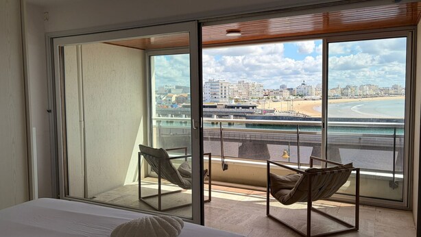 A hotel room with a large sliding glass door leading to a balcony with a view of the beach and cityscape.