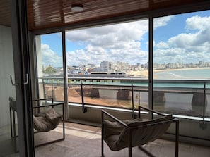 Un balcon avec deux fauteuils confortables, une vue sur une plage et un panorama sur la ville de l’autre c ôté de l’eau.