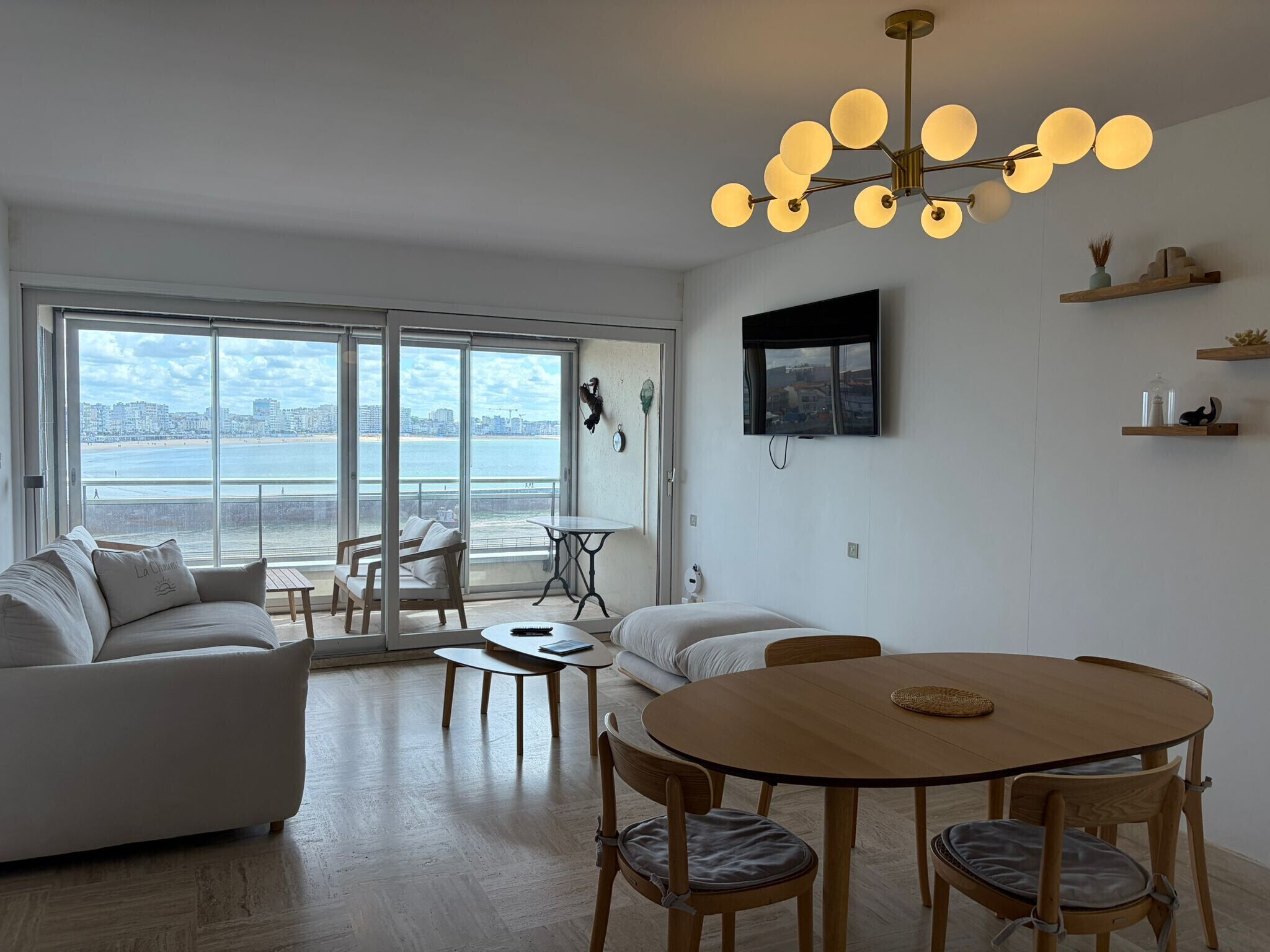 A spacious living area with a large window overlooking a beach, a round wooden dining table, and a modern chandelier.