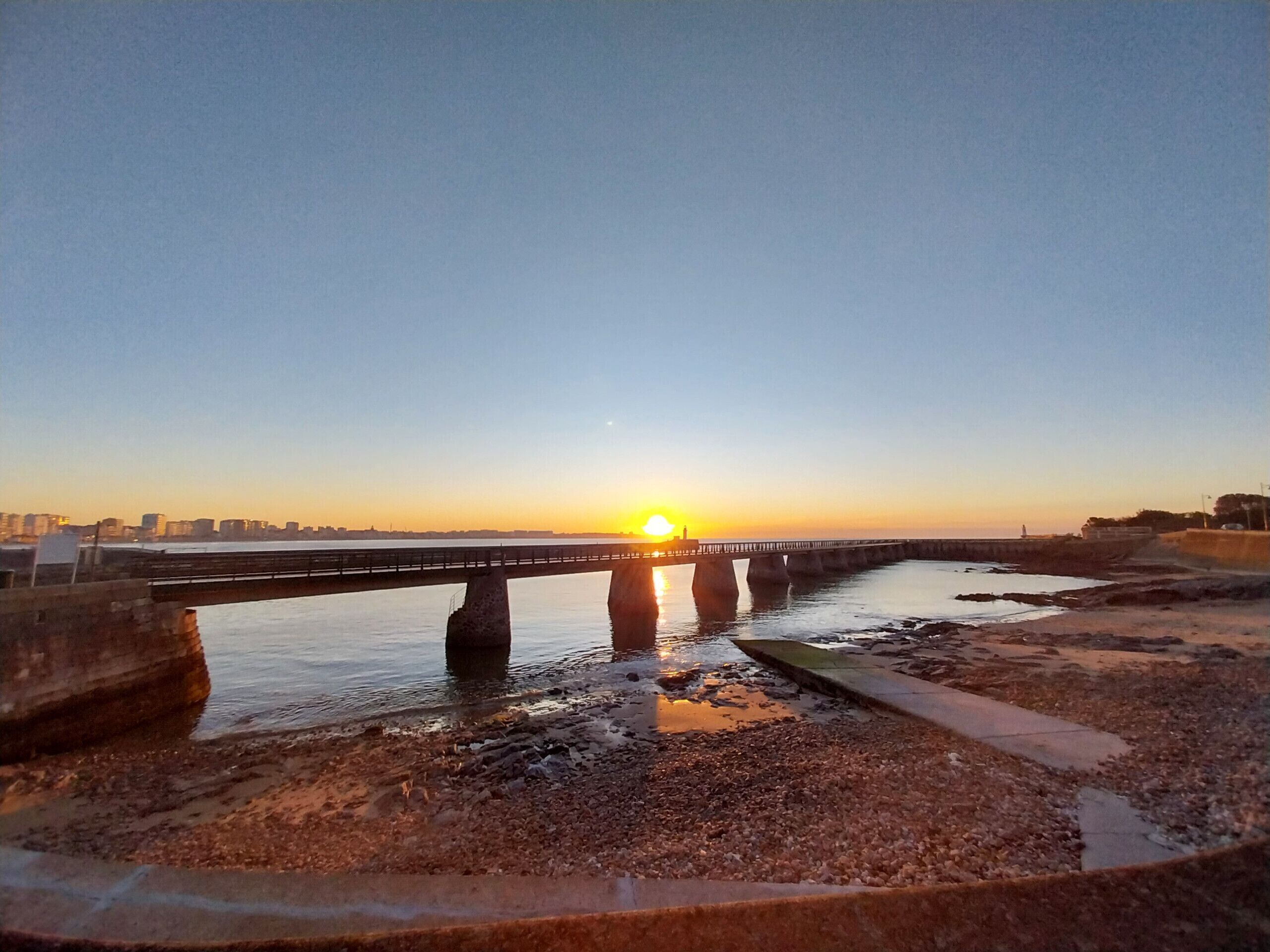 Un coucher de soleil sur une rivière, avec un pont, une rive rocheuse et les contours d’une ville lointaine.