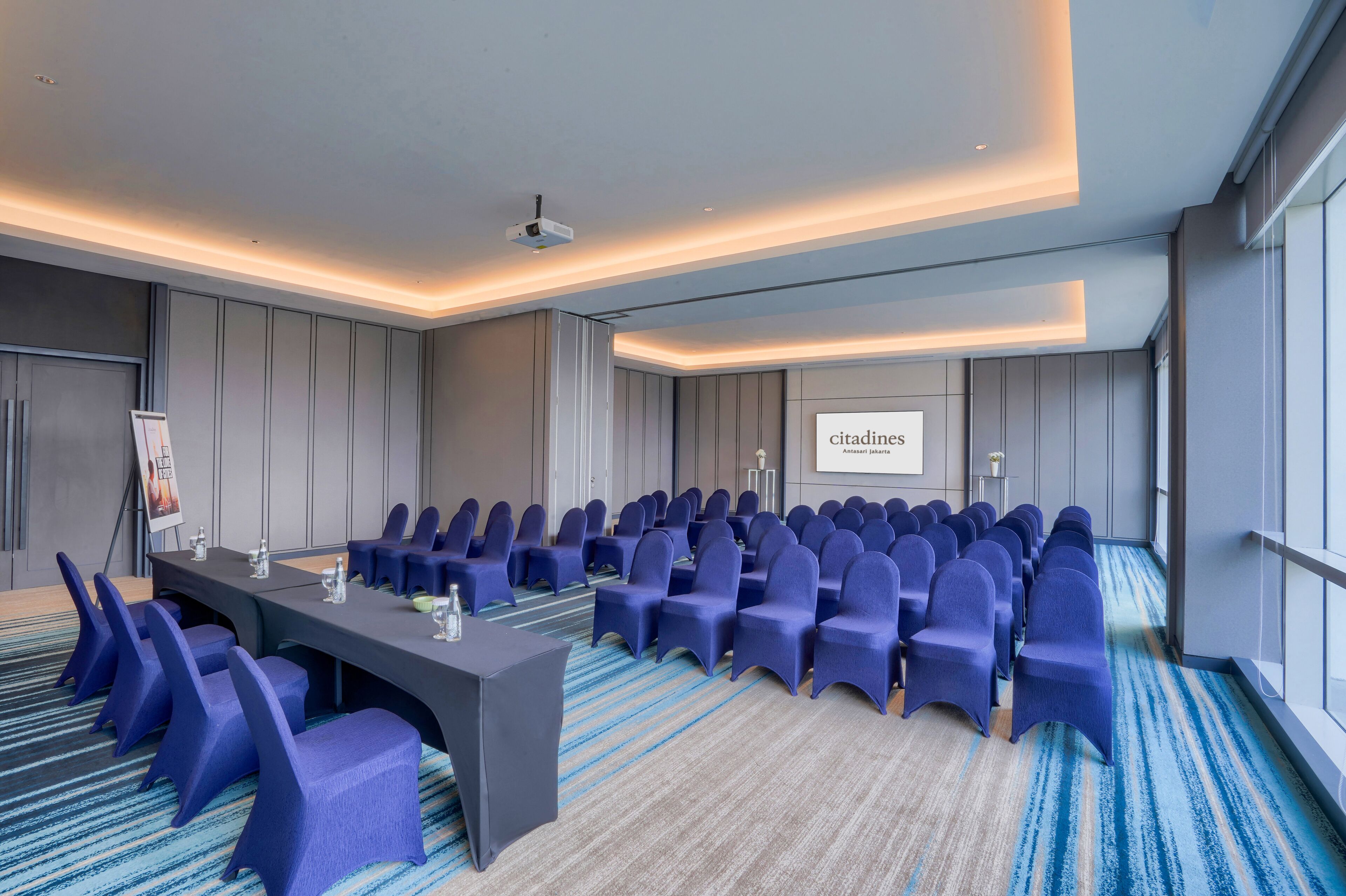 A conference room with a long table, rows of chairs, a projector screen, and a carpeted floor.
