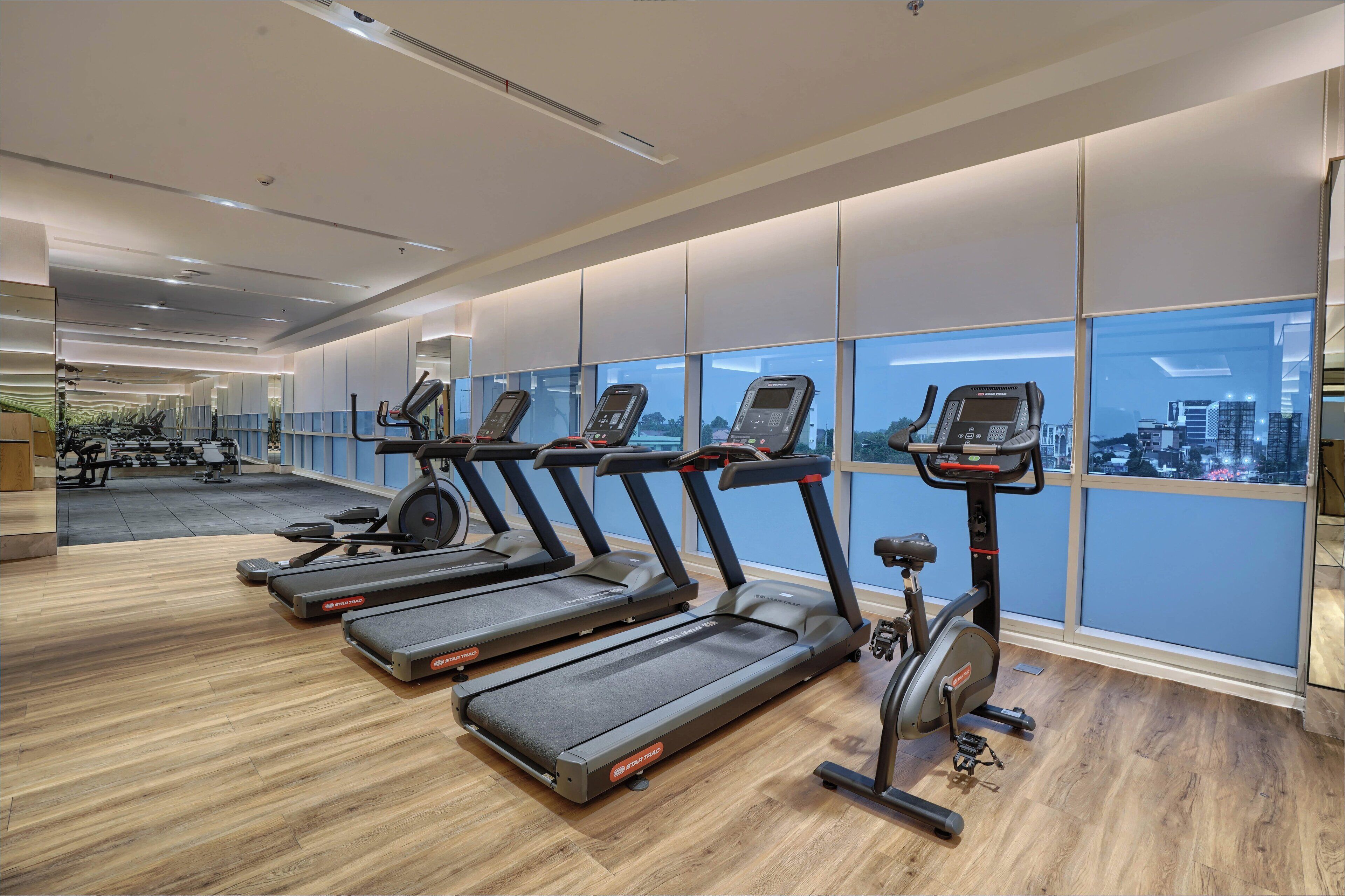Un gimnasio moderno con varias cintas de correr y una máquina elíptica, ventanas amplias con vista a la ciudad y suelo de madera.