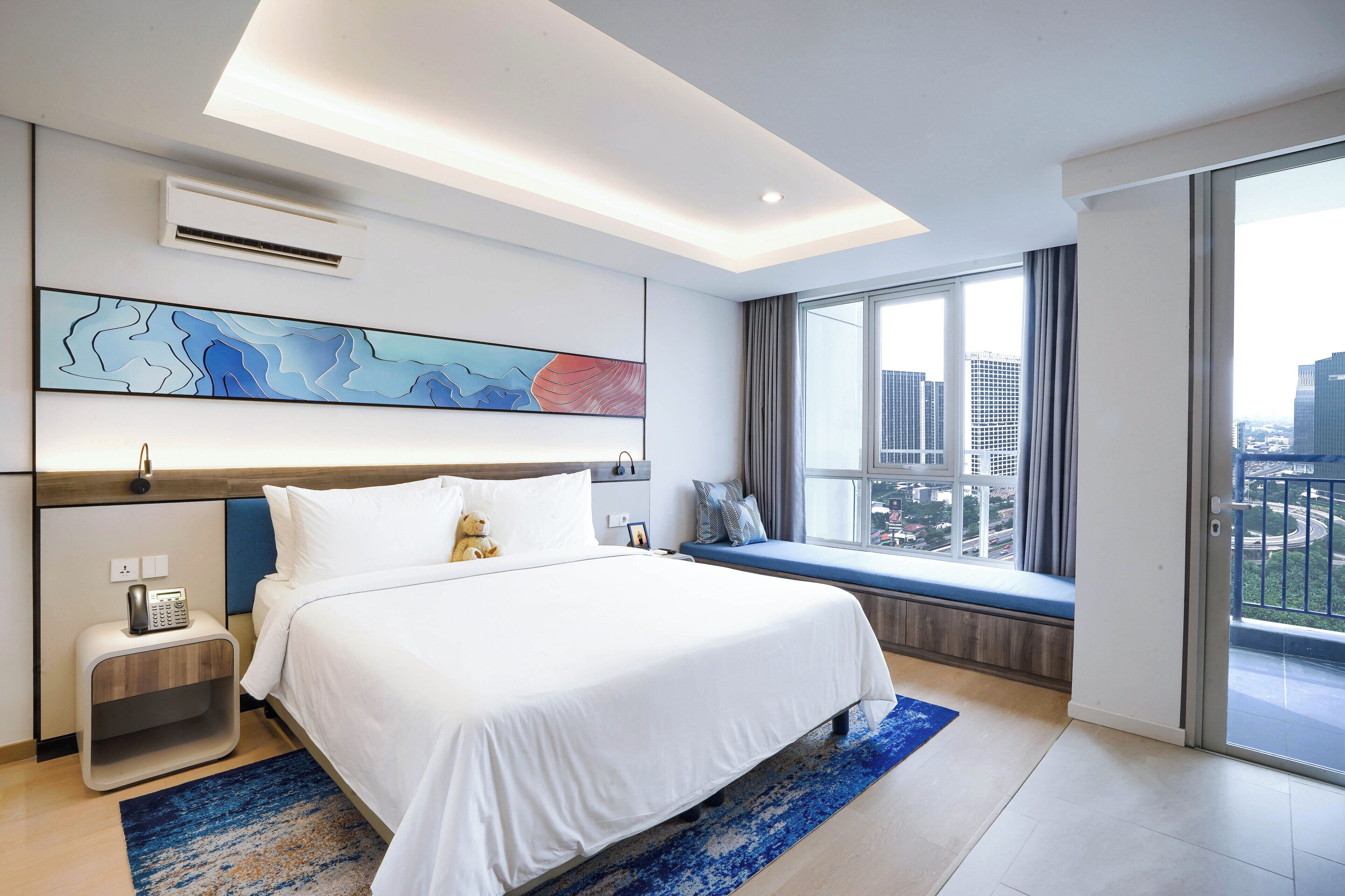 A modern hotel room with a large bed, a bedside table with a telephone, a window with a view of the city, and a ceiling light.