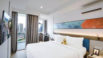 A hotel room with a large bed, a desk, a chair, a nightstand, and a view of the cityscape through the window.