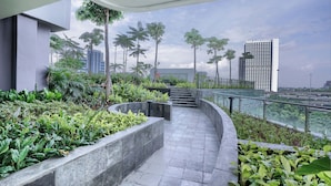 Une promenade extérieure moderne, au design incurvé, bordée de jardinières et de palmiers, qui mène à un bâtiment doté d’une balustrade en verre offrant une vue sur le paysage urbain.